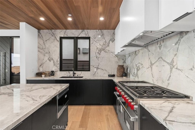 kitchen featuring wall chimney exhaust hood, dark cabinets, light stone countertops, light wood-style floors, and a sink