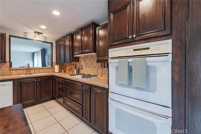 kitchen with dark brown cabinetry, white appliances, tile counters, backsplash, and light tile patterned flooring