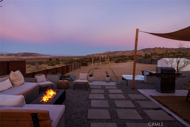 patio terrace at dusk featuring an outdoor living space with a fire pit, a fenced backyard, a grill, a mountain view, and a playground