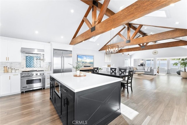 kitchen featuring open floor plan, built in appliances, light countertops, dark cabinetry, and wall chimney range hood