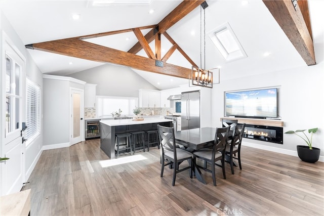 dining room with light wood finished floors, beverage cooler, a glass covered fireplace, a chandelier, and beam ceiling