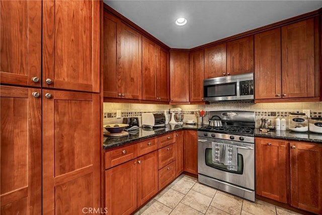 kitchen featuring backsplash, appliances with stainless steel finishes, brown cabinetry, light tile patterned flooring, and dark stone counters