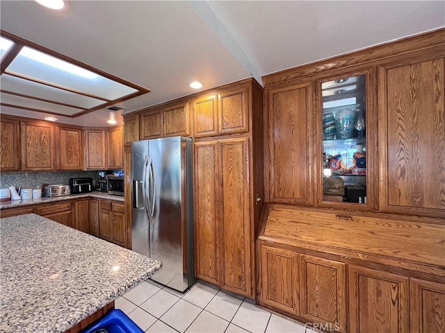 kitchen with stainless steel fridge, light stone counters, brown cabinets, and light tile patterned flooring