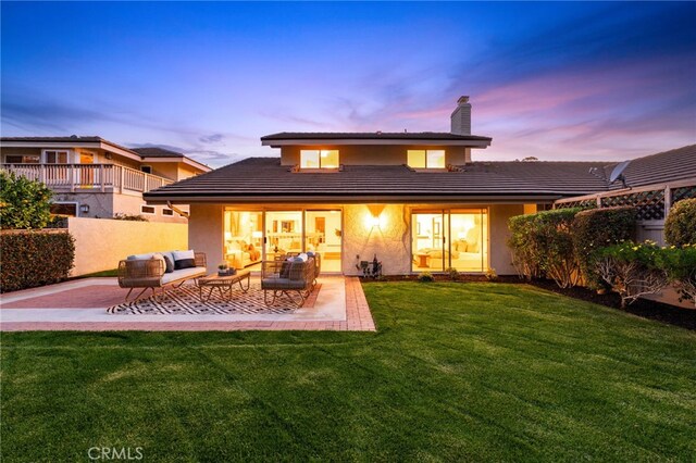 back of house at dusk featuring stucco siding, an outdoor living space, a patio, a yard, and a chimney