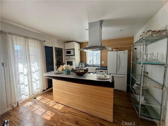 kitchen featuring island range hood, white appliances, a sink, wood finished floors, and dark countertops