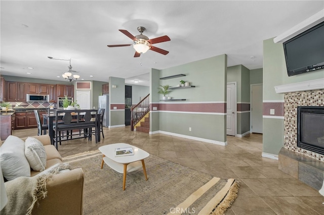 living area with light tile patterned floors, a tile fireplace, ceiling fan with notable chandelier, baseboards, and stairs