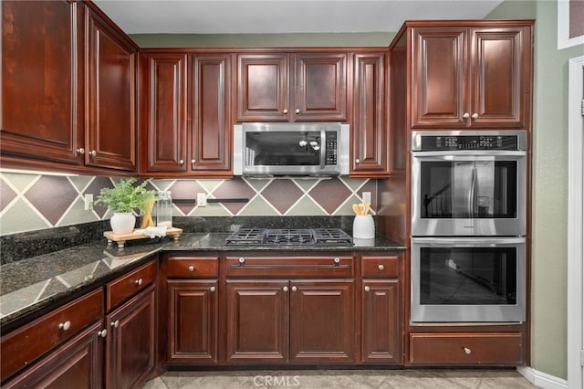 kitchen featuring reddish brown cabinets, backsplash, stainless steel appliances, and dark stone countertops