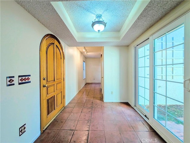 corridor with a tray ceiling, a textured ceiling, baseboards, and tile patterned floors