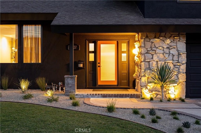 entrance to property featuring a garage, stone siding, and roof with shingles