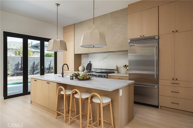 kitchen featuring light wood finished floors, tasteful backsplash, an island with sink, stainless steel built in refrigerator, and a kitchen bar