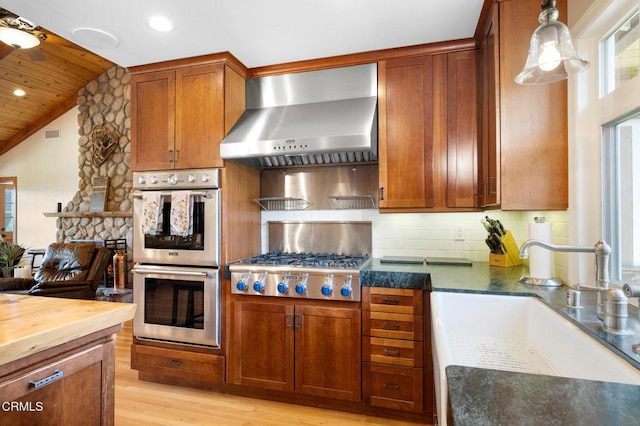 kitchen with light wood finished floors, wall chimney exhaust hood, a sink, stainless steel appliances, and backsplash