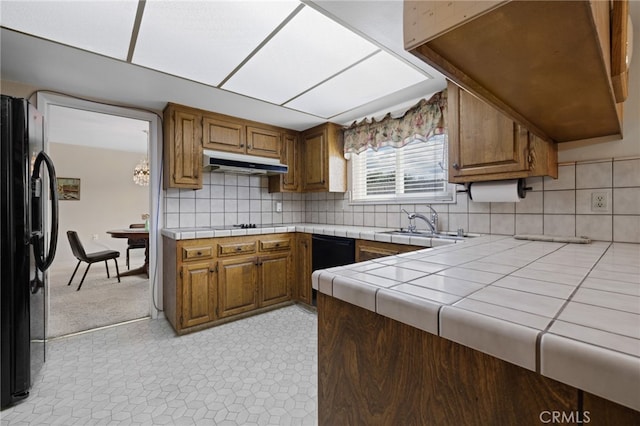 kitchen with under cabinet range hood, a sink, tile counters, black appliances, and brown cabinetry