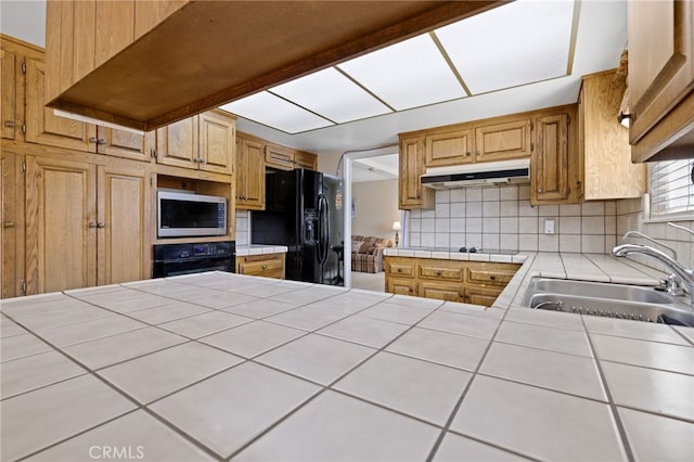 kitchen with under cabinet range hood, a sink, tile counters, decorative backsplash, and black appliances