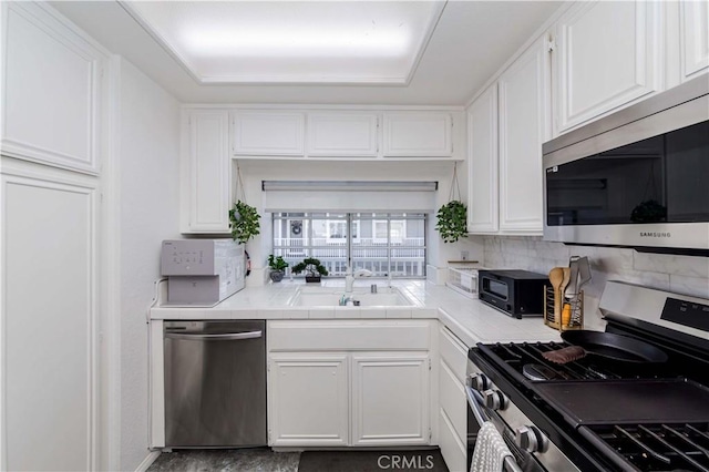 kitchen with tile countertops, stainless steel appliances, a sink, white cabinets, and backsplash