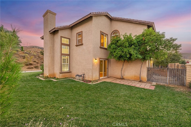 back of house at dusk with a lawn, a chimney, fence, and stucco siding