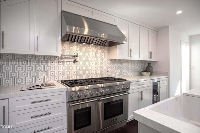 kitchen featuring backsplash, white cabinets, wall chimney range hood, light stone countertops, and double oven range