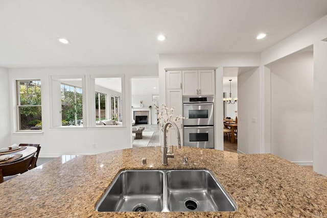 kitchen featuring a fireplace, an inviting chandelier, double oven, a sink, and light stone countertops