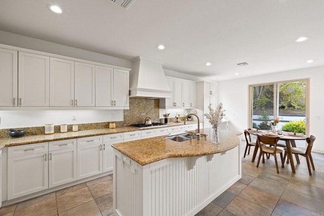 kitchen featuring a kitchen island with sink, premium range hood, a sink, decorative backsplash, and stainless steel gas stovetop