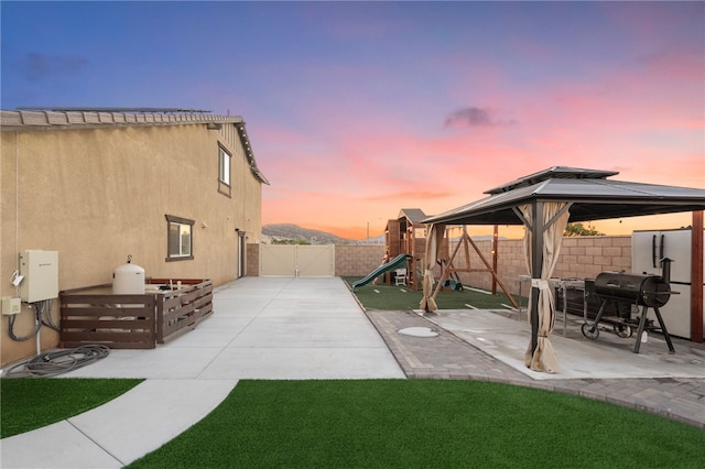 patio terrace at dusk featuring a gazebo, area for grilling, a fenced backyard, and a playground