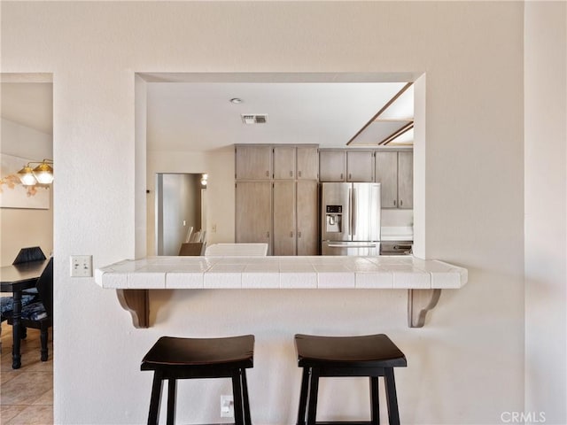 kitchen featuring visible vents, stainless steel fridge with ice dispenser, tile countertops, a breakfast bar, and a peninsula