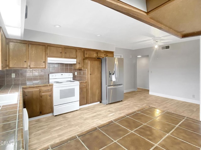 kitchen featuring tile countertops, under cabinet range hood, white electric range, backsplash, and stainless steel fridge