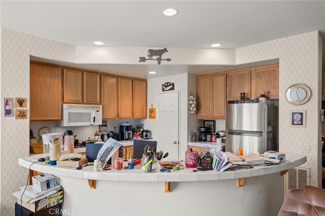 kitchen featuring white microwave, a peninsula, tile counters, freestanding refrigerator, and wallpapered walls