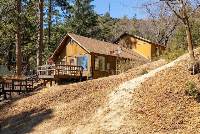 view of side of home featuring a shingled roof, a deck, and a wooded view