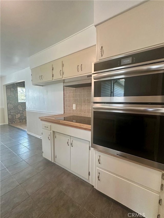 kitchen with a wainscoted wall, black electric cooktop, white cabinets, stainless steel double oven, and under cabinet range hood