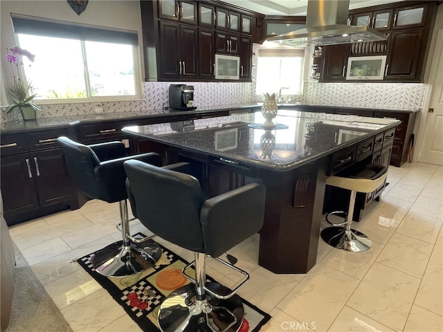 kitchen featuring marble finish floor, tasteful backsplash, stainless steel microwave, island range hood, and a kitchen breakfast bar