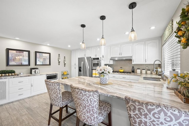 kitchen with light stone counters, a sink, stainless steel appliances, under cabinet range hood, and a kitchen breakfast bar