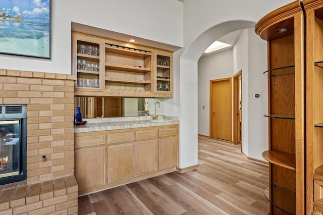 kitchen with a sink, a brick fireplace, tile counters, light wood finished floors, and glass insert cabinets