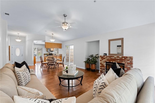 living room featuring ceiling fan, visible vents, vaulted ceiling, a brick fireplace, and light wood finished floors