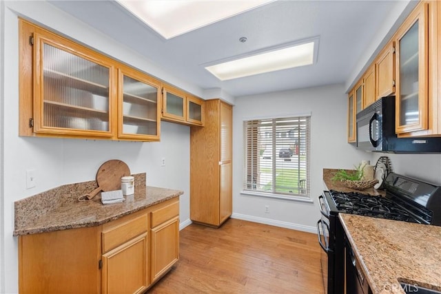 kitchen featuring baseboards, light wood-style floors, light stone countertops, black appliances, and glass insert cabinets