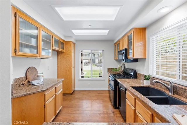 kitchen with glass insert cabinets, light stone countertops, light wood-type flooring, black appliances, and a sink