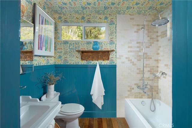 bathroom with toilet, a wainscoted wall, wood finished floors, washtub / shower combination, and wallpapered walls