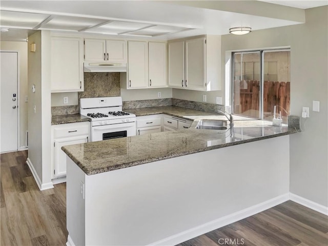 kitchen featuring white gas stove, under cabinet range hood, a peninsula, a sink, and dark stone countertops