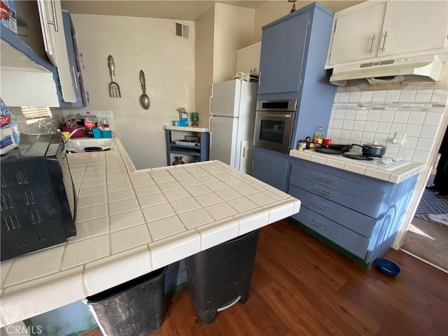 kitchen with tile countertops, blue cabinets, under cabinet range hood, white appliances, and visible vents