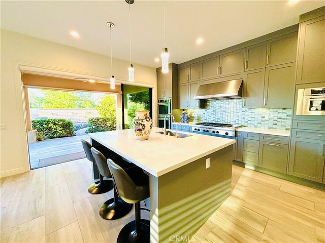 kitchen with tasteful backsplash, light countertops, a sink, an island with sink, and under cabinet range hood