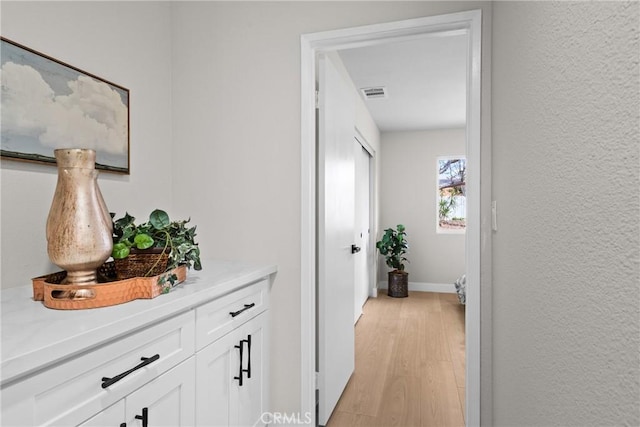 hallway featuring a textured wall, visible vents, baseboards, and light wood finished floors