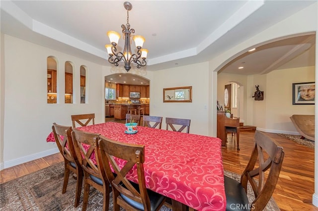 dining area with baseboards, arched walkways, a raised ceiling, wood finished floors, and an inviting chandelier