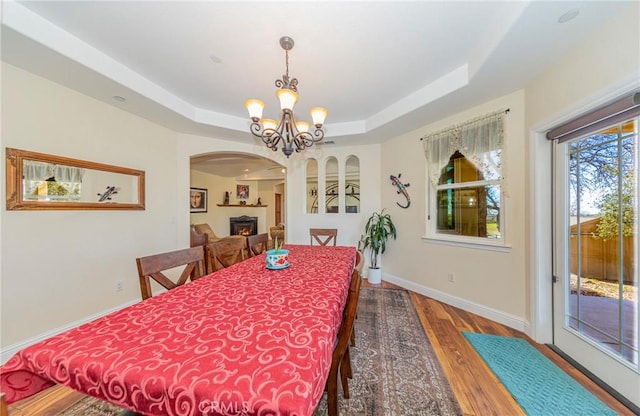 dining space with a notable chandelier, a raised ceiling, wood finished floors, a lit fireplace, and baseboards