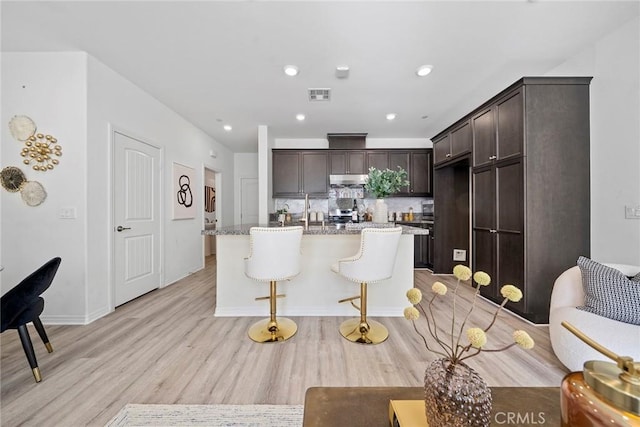 kitchen featuring under cabinet range hood, visible vents, dark brown cabinets, light wood-type flooring, and an island with sink