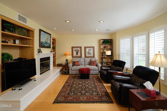 living area with built in shelves, recessed lighting, visible vents, light wood-type flooring, and a glass covered fireplace