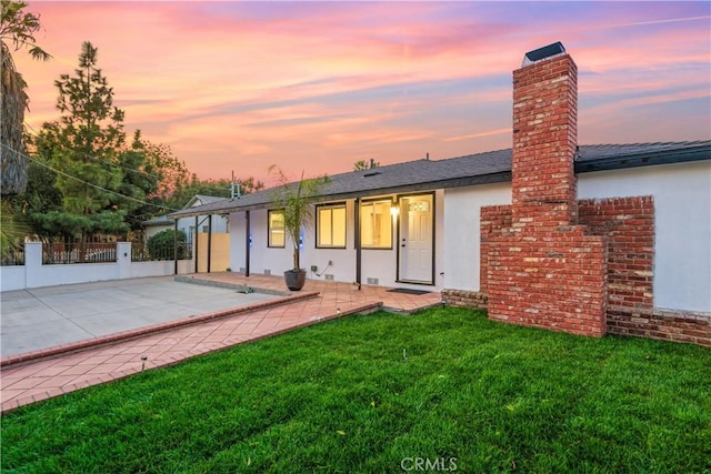 back of house at dusk featuring roof with shingles, a yard, stucco siding, a patio area, and fence