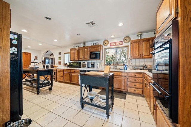 kitchen with arched walkways, visible vents, backsplash, black appliances, and brown cabinetry