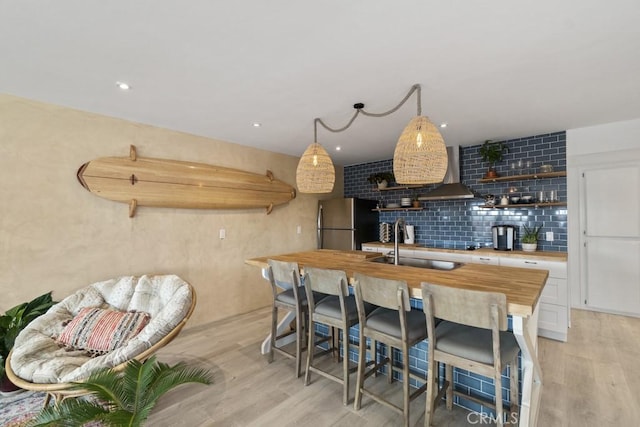 kitchen with light wood-type flooring, freestanding refrigerator, wall chimney range hood, and open shelves