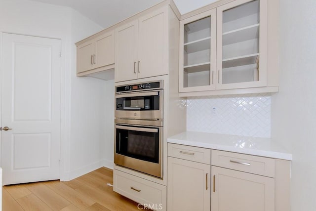 kitchen featuring tasteful backsplash, glass insert cabinets, light countertops, stainless steel double oven, and light wood-style floors