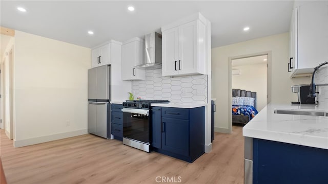 kitchen with light wood finished floors, blue cabinetry, a sink, appliances with stainless steel finishes, and wall chimney range hood