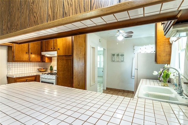 kitchen featuring under cabinet range hood, white range with gas stovetop, a sink, tile counters, and brown cabinets