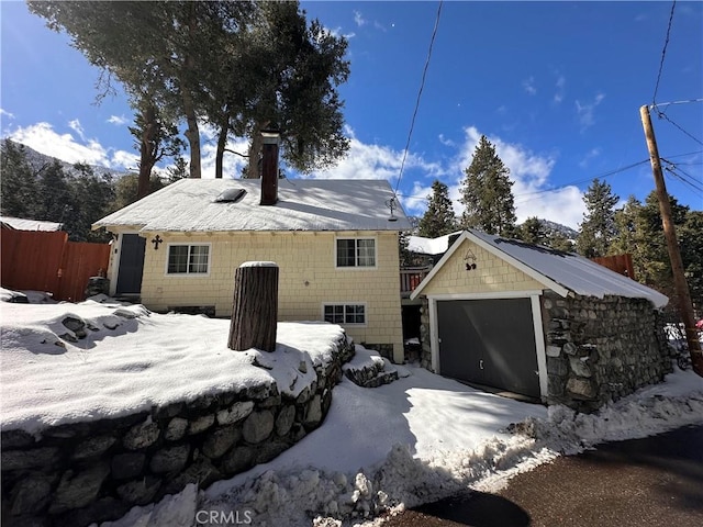 snow covered rear of property featuring a detached garage, fence, and an outbuilding